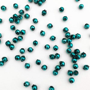 May include: A scattering of teal blue round beads on a white background. The beads are shiny and have a metallic finish.