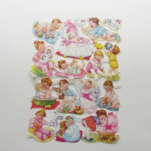 May include: A sheet of vintage baby-themed die-cut paper stickers. The stickers feature illustrations of babies in various poses, including playing with toys, in a bassinet, and being bathed. The color palette includes pastels like pink, blue, and yellow.