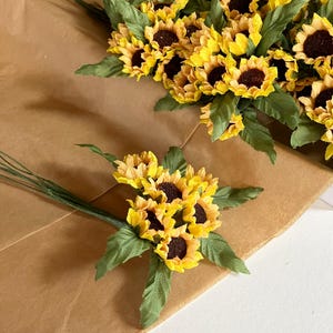 May include: Arrangement of artificial sunflowers with bright yellow petals and dark brown centers, accented by green leaves. The sunflowers are arranged on brown paper, creating a rustic and natural aesthetic. Perfect for home decor or crafting.
