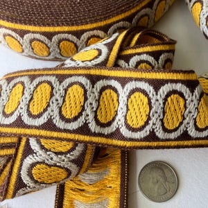 May include: A roll of woven fabric trim featuring a repeating pattern of yellow oval shapes outlined in white, set against a dark brown background. A United States quarter coin is placed next to the trim to provide a sense of scale for the width.