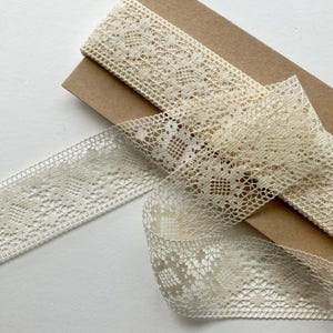 May include: Ivory-colored lace ribbon with intricate openwork patterns. The ribbon is partially rolled and laid on a brown cardboard surface. The lace has a delicate, vintage aesthetic, suitable for crafts and embellishments.