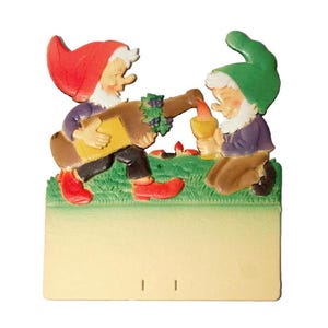 May include: A whimsical figurine featuring two gnomes. One gnome pours wine from a bottle adorned with grapes into a goblet held by the other. The gnomes wear red and green hats, purple jackets, and red shoes. The base is a light beige color.