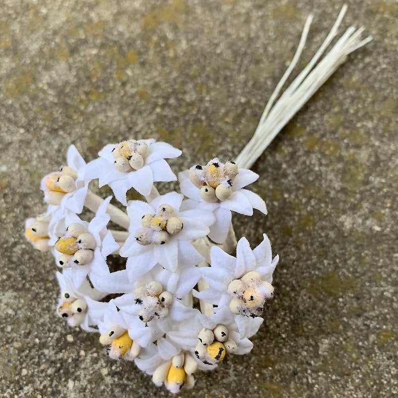 May include: A cluster of white fabric flowers with yellow and tan centers and a bundle of off-white stems. The flowers have a star-like shape and are arranged in a small bouquet. The background is a textured, light gray surface.