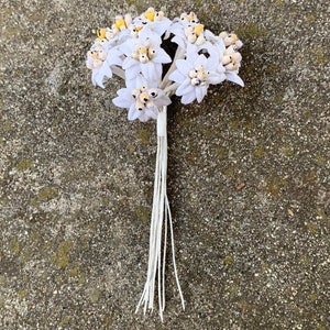 May include: A bouquet of white artificial flowers with yellow centers and a bundle of white string stems. The flowers are clustered together, and the stems are tied with white tape. The background is a textured, gray surface.