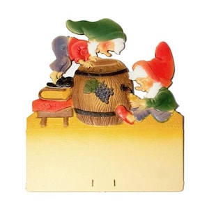 May include: Two gnomes, one in green and one in red, are looking into a wooden barrel with a bunch of grapes carved on the side. The gnomes are sitting on a yellow surface with a red and white striped surface behind them. There is a stack of books and a small wooden table in the background.