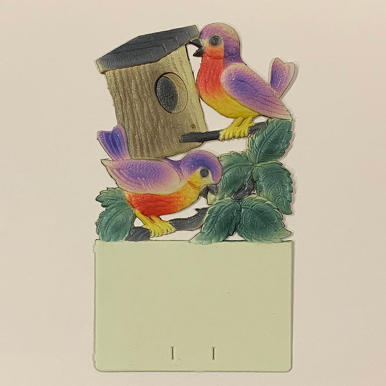 May include: A vintage paper craft featuring two purple and yellow birds perched on a green leafy branch. The birds are near a small wooden birdhouse with a round opening. The craft is attached to a light green card with the number "11" printed on it.