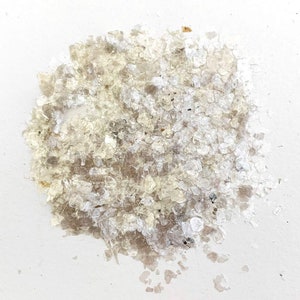 May include: A pile of translucent, iridescent mica flakes. The flakes vary in size and shape, with a mix of clear, white, and light brown tones. The mica has a shimmering, reflective quality, suggesting its use in crafts or cosmetics.
