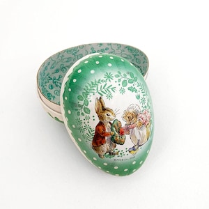 May include: A green and white Easter egg-shaped tin with a polka dot pattern. The tin features a picture of Peter Rabbit and a hedgehog. The text "© F.W.& Co. 2023" is printed on the bottom of the tin.