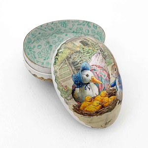 May include: An egg-shaped decorative box with a hinged lid. The box features a detailed illustration of a duck in a blue bonnet with ducklings, a rabbit, and a hen. The interior of the lid has a green floral pattern.