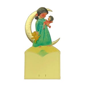 May include: A girl doll in a green dress holds a smaller doll in a red dress. The dolls are standing on a crescent moon with a gold finish. The moon is attached to a yellow card with a blank space for writing.