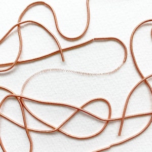 May include: A close-up of a tangled pile of copper-colored elastic cord on a white background. The cord is thin and flexible, and it is arranged in a random pattern.