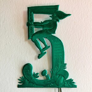 May include: A metallic green decorative figure of a person climbing a frame, atop a stylized number 5. The figure is detailed with a hat and shoes. The base of the number 5 has floral and plant-like embellishments.