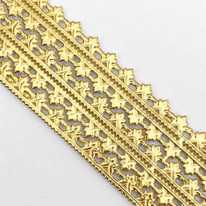 May include: Close-up of a gold-colored decorative trim with an ornate, repeating floral and leaf pattern. The trim has a scalloped edge and is made of a shiny, metallic material. The design is symmetrical and detailed.