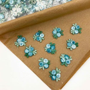 May include: Close-up of several small, embroidered flower appliques. Each applique features a cluster of blue and teal flowers with green leaves. The appliques are arranged on a brown paper surface, with more in the background.