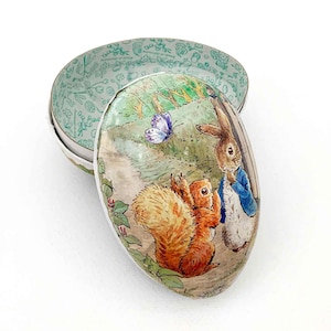 May include: An oval-shaped decorative tin with a hinged lid. The lid features a painted scene of a squirrel and a rabbit in a forest setting, with a butterfly. The interior of the tin is light green with a floral pattern.