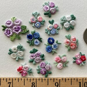 May include: A collection of thirteen small, embroidered floral appliques featuring various color combinations of pink, purple, blue, white, and red thread with green leaves. The embroidered flowers are arranged on a surface above a wooden ruler.
