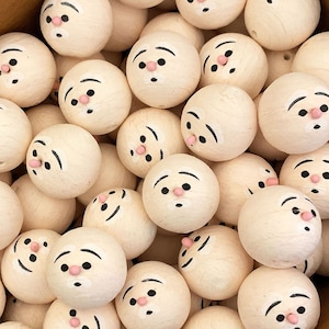 May include: A collection of round, light-colored wooden beads, each hand-painted with a whimsical face. The faces feature black eyebrows, eyes, and a small pink nose and mouth. The beads are closely packed together.