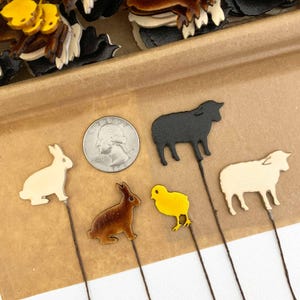 May include: A collection of farm animal-themed craft embellishments. The image shows a black sheep, a white sheep, a yellow chick, and two rabbits in cream and brown. A US quarter is included for size reference. The embellishments are on thin brown sticks.