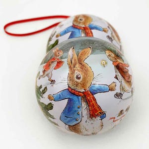 May include: Two round Christmas ornaments featuring a winter scene with Peter Rabbit and friends ice skating. The ornaments are white with a silver metallic finish and have a red ribbon for hanging.