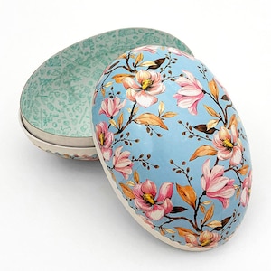 May include: An egg-shaped decorative box with a light blue exterior adorned with pink and yellow floral branches. The interior of the box is light green with a pattern of small shapes. The box is open, revealing the interior.