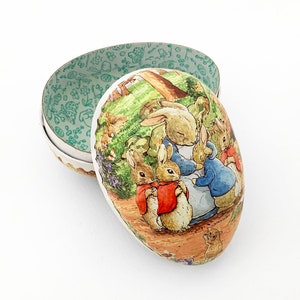 May include: An open, egg-shaped container with a lid featuring a scene of rabbits in a garden. The interior of the container is light green with a floral pattern. The exterior has a cream-colored border.