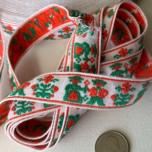 May include: A roll of woven fabric trim featuring a repeating pattern of figures in green and orange on a white background. The trim is displayed next to a United States quarter coin for scale, showing the width of the decorative textile ribbon.
