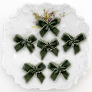 May include: Six olive green velvet bows arranged on a white, scalloped-edge plate with floral accents. The bows are tied with a classic bow shape, and the velvet has a soft, textured appearance. The bows are a craft supply.