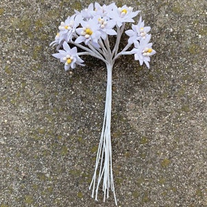 K&ouml;nnte beinhalten: Ein Bund k&uuml;nstlicher wei&szlig;er Blumen mit gelben Zentren. Die Blumen haben mehrere Bl&uuml;tenbl&auml;tter und sind auf einem grauen Stiel angeordnet. Die Stiele sind mit wei&szlig;er Schnur zusammengebunden.