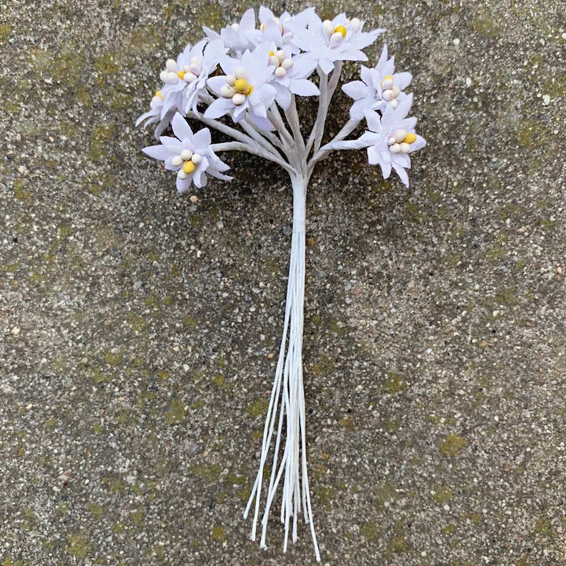 K&ouml;nnte beinhalten: Ein Bund k&uuml;nstlicher wei&szlig;er Blumen mit gelben Zentren. Die Blumen haben mehrere Bl&uuml;tenbl&auml;tter und sind auf einem grauen Stiel angeordnet. Die Stiele sind mit wei&szlig;er Schnur zusammengebunden.