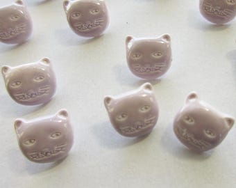 May include: A collection of ten lavender-colored ceramic buttons shaped like cat faces. Each button has a white raised design of a cat's face with eyes, nose, and whiskers.