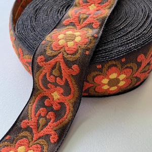 May include: A roll of dark brown woven trim featuring a repeating embroidered pattern of orange and yellow flowers with brown centers. The decorative ribbon has a textured fabric surface and a continuous floral design that wraps around the spool.