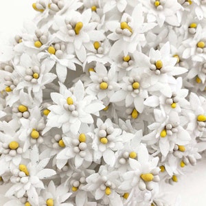K&ouml;nnte beinhalten: Eine Ansammlung kleiner, wei&szlig;er Papierblumen mit gelben und grauen Zentren. Die Blumen haben mehrere Bl&uuml;tenbl&auml;tter und sind dicht gepackt, wodurch eine strukturierte, florale Anordnung entsteht. Der Gesamteffekt ist zart und detailliert.