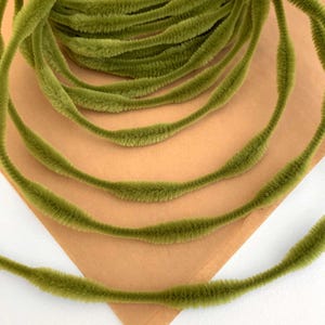 May include: Close-up of olive green chenille stems, arranged in a spiral, resting on a tan surface. The stems have a soft, fuzzy texture and are ideal for craft projects. The image shows the stems in a close-up view.