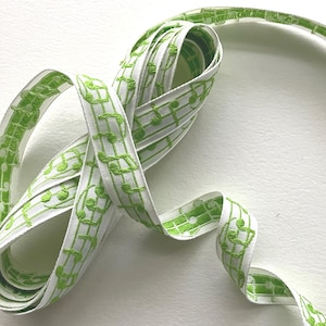 May include: White ribbon with a repeating pattern of green musical notes. The ribbon is curled and appears to be made of fabric. The musical notes are embroidered onto the ribbon.