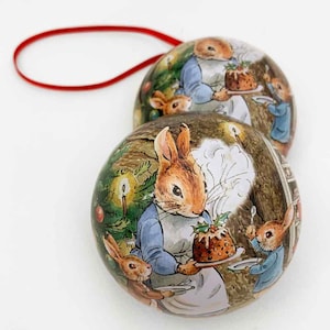 May include: Two round Christmas ornaments featuring a whimsical illustration of bunnies in a cozy winter scene. The ornaments depict bunnies wearing blue and white clothing, holding a Christmas pudding and a plate. The ornaments are decorated with a brown background and a green Christmas tree.