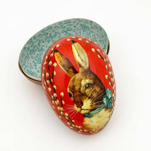 May include: A red, egg-shaped decorative box with a rabbit illustration. The rabbit has brown fur and a blue bow. The box is open, revealing a teal interior with a floral pattern. The box is decorated with white pussy willow branches.