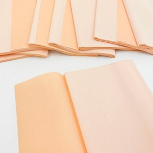 May include: A collection of folded tissue paper sheets in shades of peach and light pink. The paper has a textured, crepe-like appearance, with some sheets partially unfolded, revealing the layered construction. Ideal for crafting or gift wrapping.