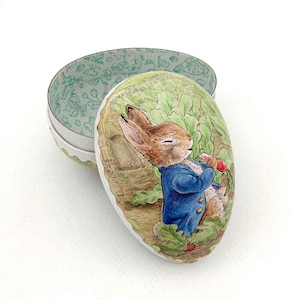 May include: An oval-shaped tin box with a lid featuring a watercolor illustration of Peter Rabbit in a blue jacket, holding a red radish. The box's interior has a green floral pattern, and the exterior is white with a green trim.