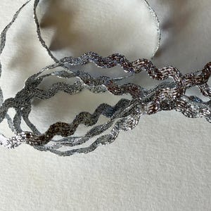 May include: Silver metallic ribbon with a wavy, textured design. The ribbon is curled and layered, creating a decorative effect. The ribbon is on a white background.