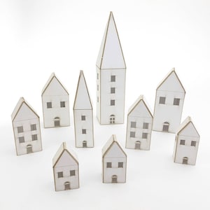 May include: A set of nine white paper model houses, including a tall church, arranged on a white surface.