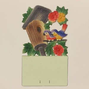 May include: A wooden birdhouse with a green roof and a brown base, surrounded by green leaves and red and yellow flowers. Two blue birds are perched on a branch in front of the birdhouse.