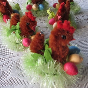 May include: Close-up of several Easter decorations. Each decoration features a brown chicken with a red comb, surrounded by green tinsel, colorful eggs, and green leaves. The decorations are arranged on a white lace tablecloth.