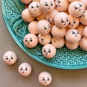 May include: A bowl filled with wooden beads, each painted a light pink color and featuring a hand-painted face with black eyes and a small mouth. The beads are arranged in a random pattern, creating a whimsical and charming display.