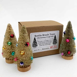 May include: A Christmas craft kit with three bottle brush trees in gold and green, adorned with colorful glass beads. The kit includes a cardboard box with a label that reads "Bottle Brush Trees Christmas Craft Kit."