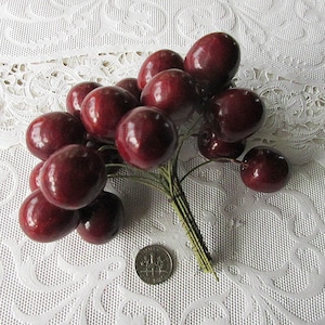May include: A bunch of artificial red cherries with green stems, arranged on a white lace tablecloth.