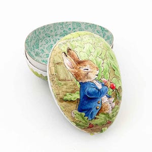 May include: An oval-shaped decorative box with a lid, featuring a watercolor illustration of Peter Rabbit in a blue jacket holding a radish. The box is open, revealing a green patterned interior. The exterior has a green and white border.