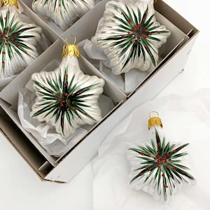 May include: White star-shaped Christmas ornaments with green and red accents are displayed in a box. Each ornament has a gold-colored cap. The ornaments are in a white box with dividers and are resting on white tissue paper.