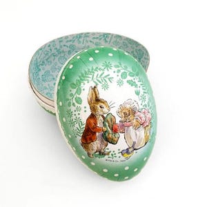 May include: An egg-shaped tin with a green lid featuring a floral and polka dot design. The lid has an illustration of a rabbit and a hedgehog. The inside of the tin is light blue with a floral pattern. The tin is open, revealing the interior.