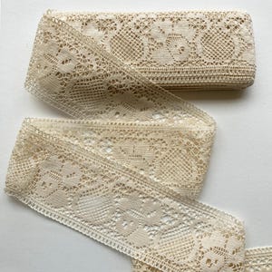 May include: A roll of antique-style beige lace ribbon. The lace features a repeating floral pattern with a delicate, open weave. The ribbon is partially unrolled, revealing its intricate design and texture, perfect for crafts and sewing projects.