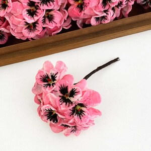 May include: A small cluster of artificial pink flowers with velvet-like petals and dark purple centers. Each flower features a central stamen with a tiny green tip. The cluster is attached to a thin, dark brown stem extending from the base of the blooms.